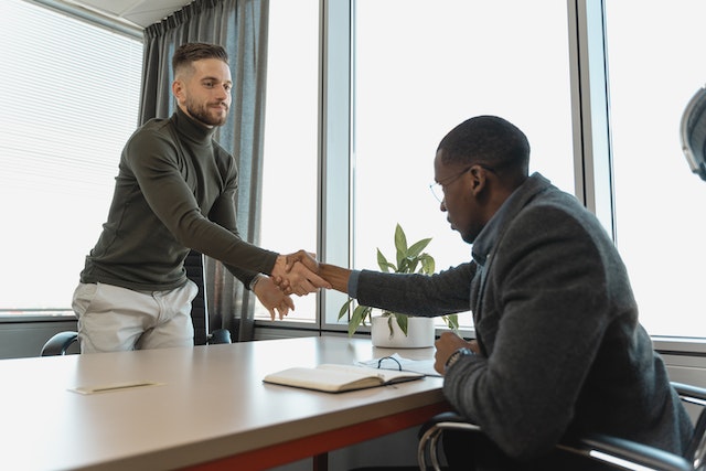 two people shaking hands across a desk in a office with large windows